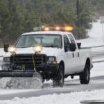 A pickup with a yellow light bar and a plow attachment clears a road of snow, with pine trees in the background.